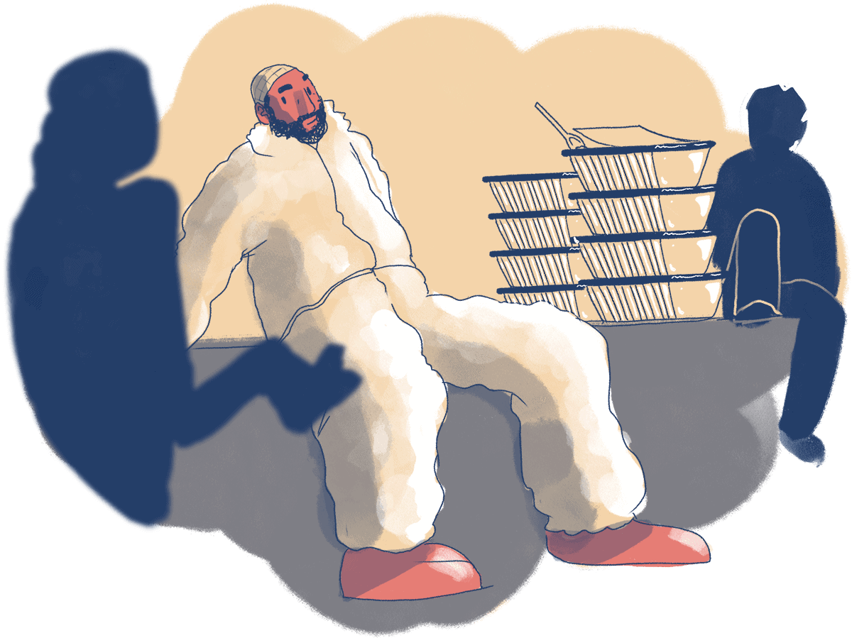 A man with a beard and skullcap is sitting down. He is wearing personal protective equipment. To his right there is a tall pile of food boxes. The one on top is open, and has a spoon in it. There are two dark blue silhouettes, one standing to his left, and one sitting to hist right